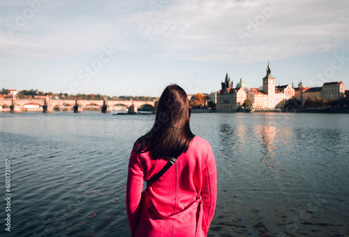Female traveler dressed in purple coat standing near the Vltava river in Prague (Czech Republic) and looking on Charles Bridge on sunset - photo from her back. Wide- angle shot of woman turist in city