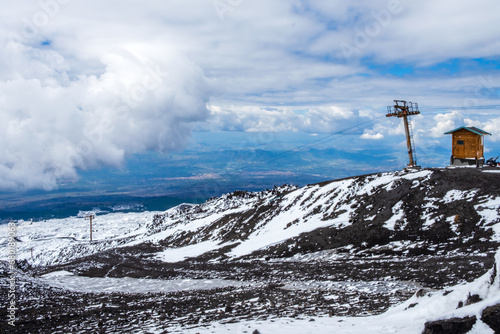 Mount Etna national park in winter. View from volcano crater with black volcanic lava stones and snow under cloudy sky and smoke. Cable car and view over Sicily island, Italy photo