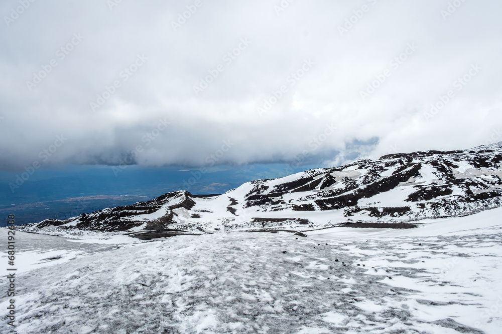 Mount Etna national park in winter. View from volcano crater with black volcanic lava stones and snow under cloudy sky and smoke. View over Sicily island, Italy