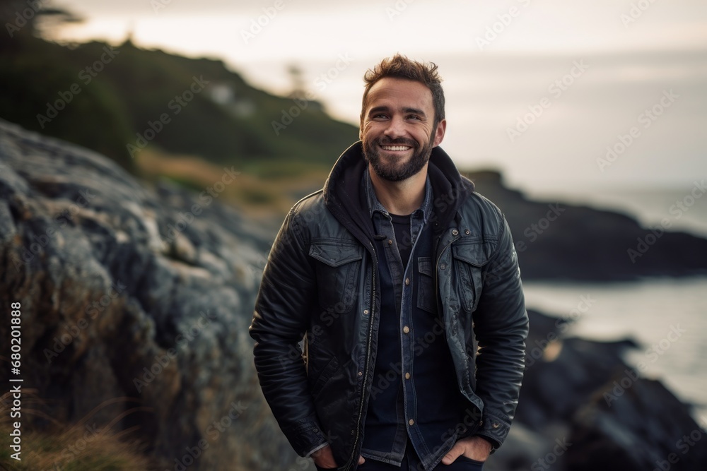 Portrait of a joyful man in his 30s sporting a stylish varsity jacket against a rocky shoreline background. AI Generation