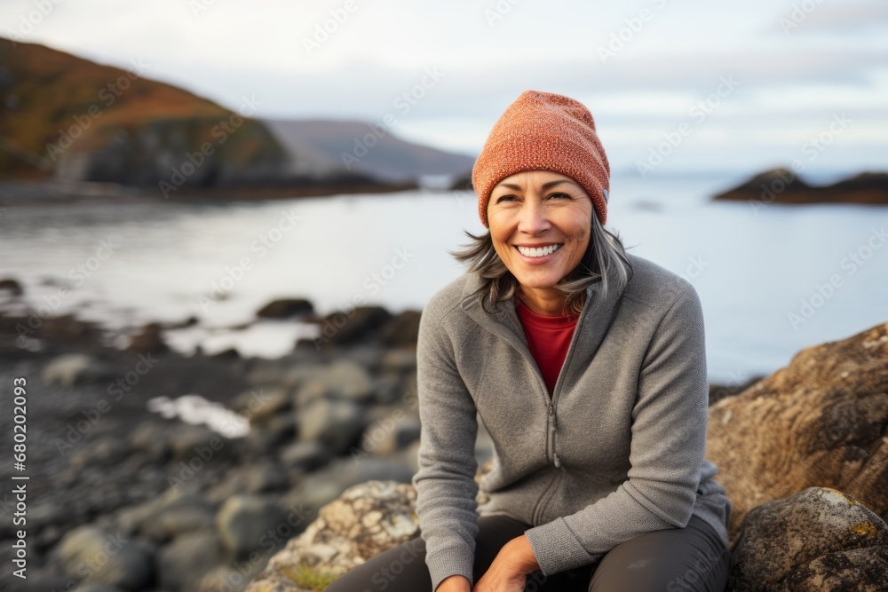 Portrait of a smiling woman in her 50s sporting a trendy beanie against a rocky shoreline background. AI Generation