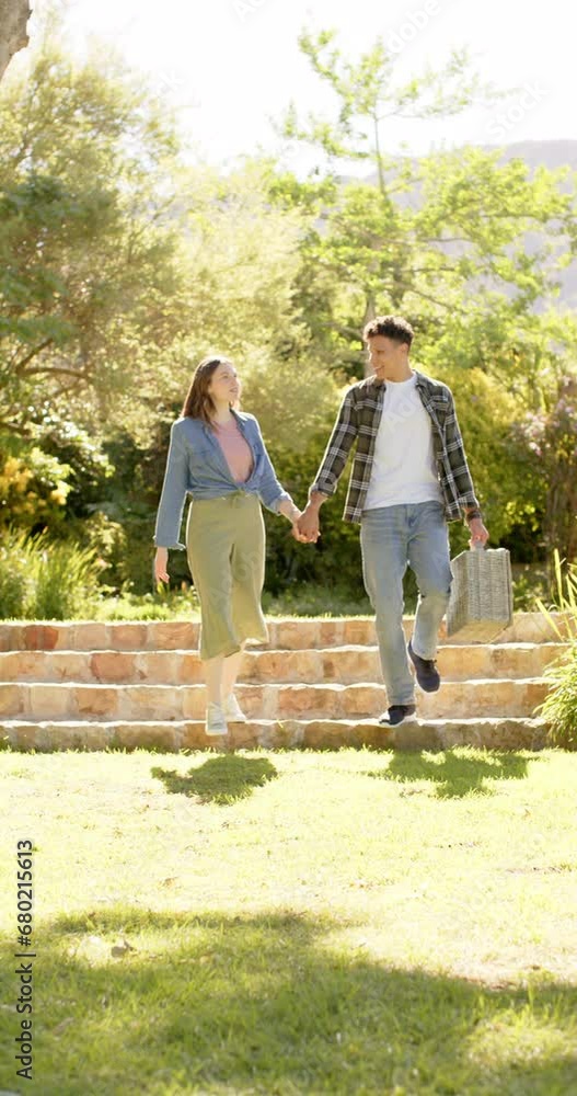 Vertical video of happy diverse couple holding hands and walking in sunny garden, in slow motion