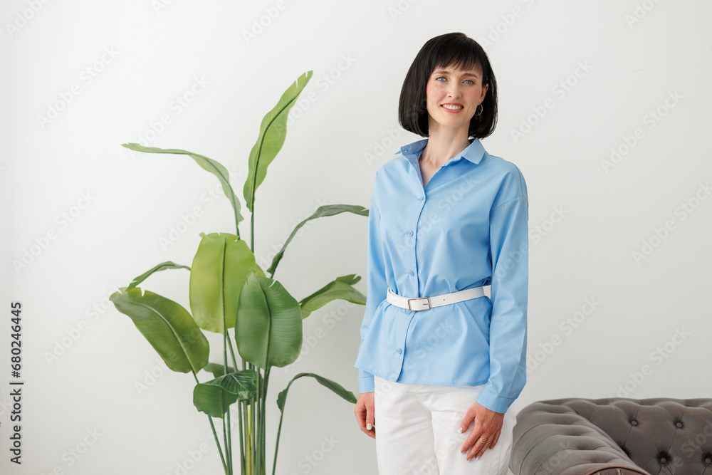 Women equality day. Smiling successful businesswoman in elegant blue shirt and white pants looks at camera at modern comfortable light office with green plant and sofa. Work life balance concept. Women equality day. Smiling successful businesswoman in elegant blue shirt and white pants looks at camera at modern comfortable light office with green plant and sofa. Work life balance concept.