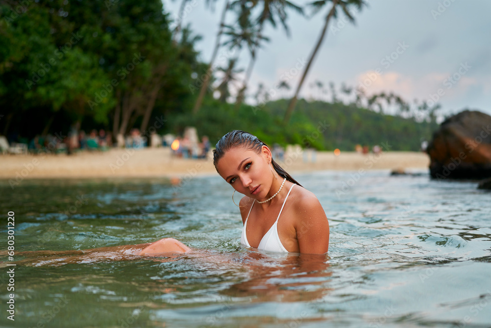 Woman enjoys swimming in ocean with durable waterproof makeup. Female in trendy swimwear relaxes in tropical sea. Traveler bathes near beach, palm trees in background, fashion, leisure concept.