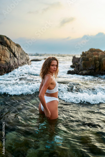 Woman in white bikini poses in sea during sunset. Female beauty fashion on rocky beach water. Summer travel photoshoot. Model on vacation by ocean.