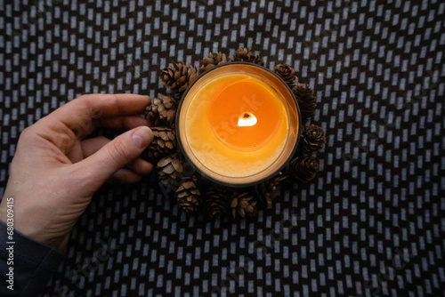 Yellow round candle in a woman's hand on a dark ornamental background flatly top view. Home decor details. Woman holding a scented aromatic burning candle among pine cones. Touch a fire. Warm light.