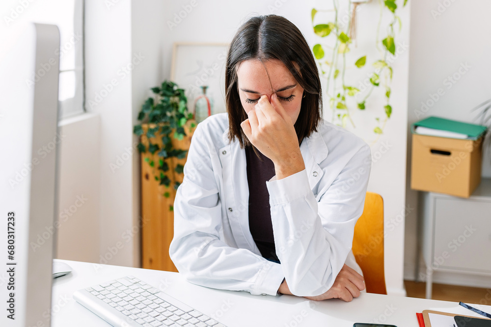 Female doctor feeling tired after working to many hours in the hospital. Frustrated professional healthcare worker with fatigue, migraine pain or anxiety sitting at workplace. Female doctor feeling tired after working to many hours in the hospital. Frustrated professional healthcare worker with fatigue, migraine pain or anxiety sitting at workplace.