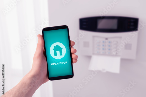 Man operating home alarm system via mobile phone against white wall with security control panel, closeup. Application interface on device screen