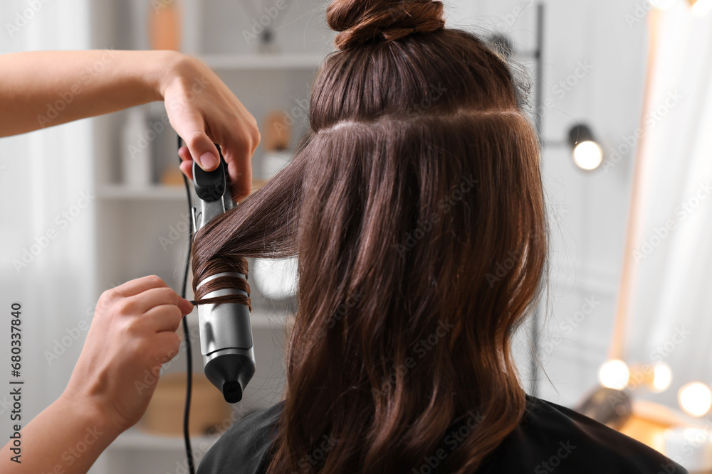 Fototapeta premium Hairdresser using curling hair iron while working with woman in salon, closeup