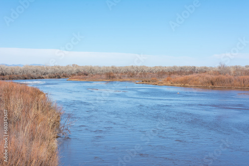 A river lined with trees and bushes in winter, with blue sky.
