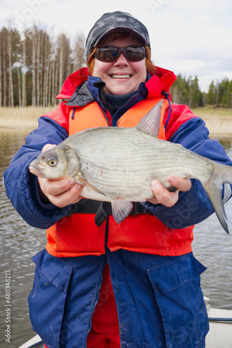 Картината върху платно A joyful woman fisherman standing by the river, holding a big bream in her hands, showcasing her successful catch and love for fishing in a beautiful natural setting