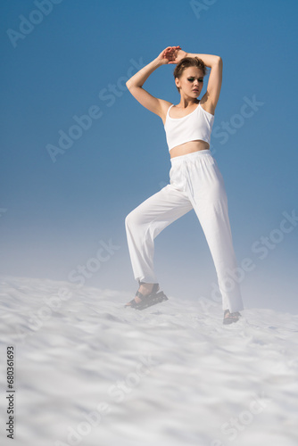 Fashionable adult woman posing hands up, eyes closed, standing through snow with light fog on sunny day with blue sky. Blond fashion model dressed in white crop top, white pants, sandals. Full length