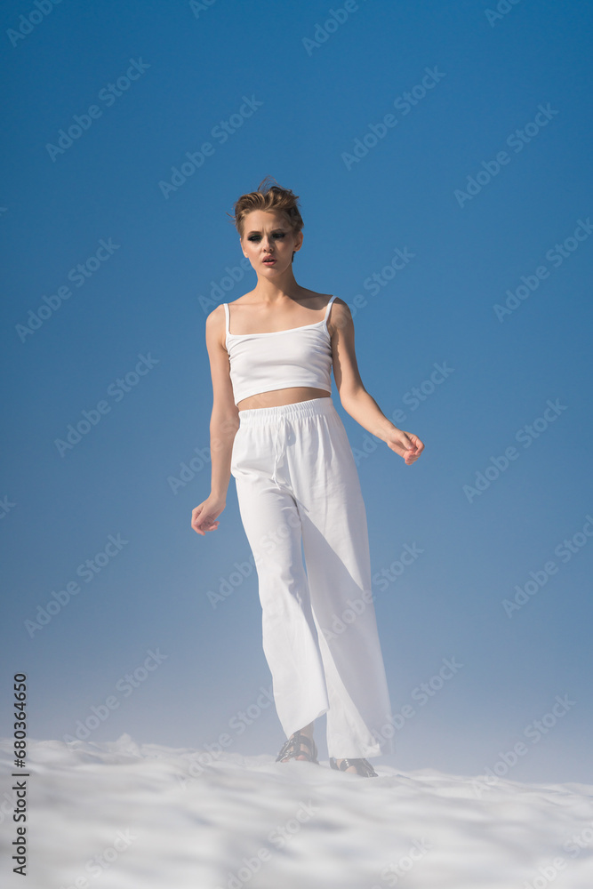 Fashionable blond woman walks through snow with light fog on sunny weather with blue sky. Authenticity adult female dressed in white cropped top, white pants and sandals. Full length, front view Fashionable blond woman walks through snow with light fog on sunny weather with blue sky. Authenticity adult female dressed in white cropped top, white pants and sandals. Full length, front view