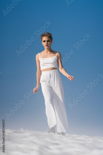Fashionable blond woman walks through snow with light fog on sunny weather with blue sky. Authenticity adult female dressed in white cropped top, white pants and sandals. Full length, front view