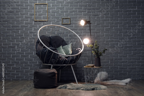 Stylish armchair and glowing lamps on table near grey brick wall