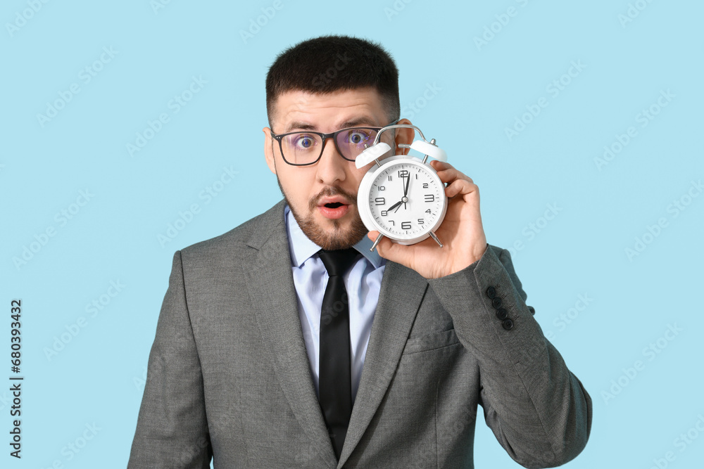 Young shocked businessman with alarm clock on blue background. Time management concept Young shocked businessman with alarm clock on blue background. Time management concept