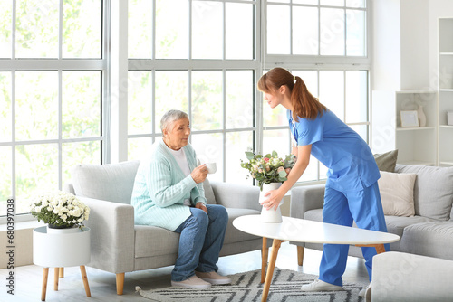 Senior woman and caregiver putting bouquet of flowers on table at home