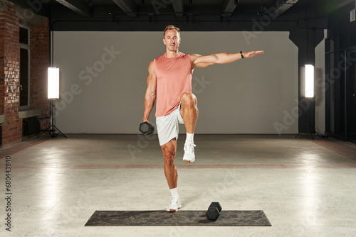 a young Caucasian athlete stands on one leg, moves his arm to the side and raises his leg, and holds a large dumbbell in his other hand. fitness. aerobics. exercises on the mat. physical health
