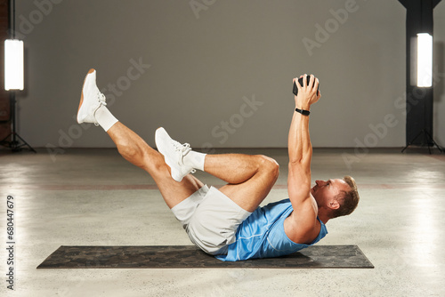 a pumped-up Caucasian athlete does a bicycle with his legs, pumping up his abs; he holds a weight in his hands, stretching them up. fitness. aerobics. exercises on the mat. physical health photo