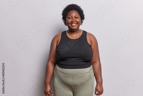 Indoor shot of dark skinned overweight woman smiles happily satisfied after workout at gym dressed in black top and leggings isolated on white background. Curly haired African female goes in for sport