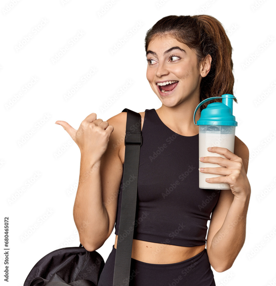 Athletic Caucasian woman holding protein shake points with thumb finger away, laughing and carefree.