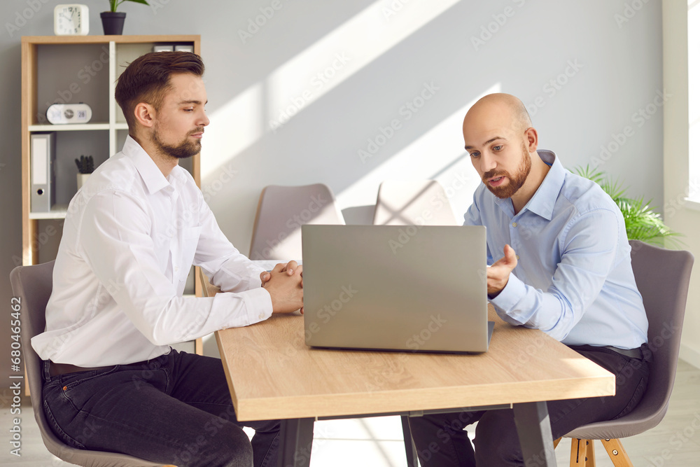 Two business men sitting at the desk on workplace talking and discussing company finance on a laptop in office. Male employers considering new projects, corporate development. Team work concept. Two business men sitting at the desk on workplace talking and discussing company finance on a laptop in office. Male employers considering new projects, corporate development. Team work concept.