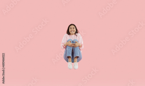 Happy excited woman doing squat jump in air. Joyful brunette girl wearing sweatshirt and jeans happily jumping hugging her knees over isolated background. Freedom, carelessness, success concept photo
