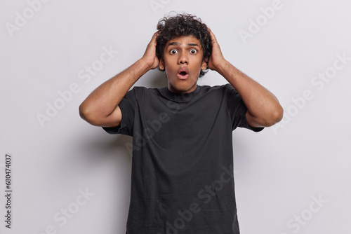 Waist up shot of scared frightened Hindu man with curly hair keeps hands on head stares amazed keeps eyes and mouth widely opened wears black t shirt isolated over white background. Omg concept photo