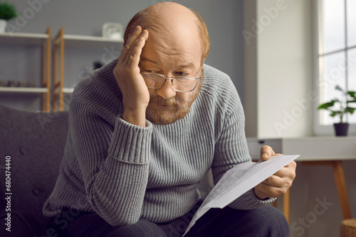 Bad news in mail. Sad, depressed senior man sitting on a chair at home, holding a last notice letter from the bank, holding his head, and thinking about finance problems of elderly people