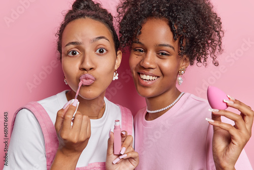 Beauty procedures and cosmetology concept. Two pretty female friends apply makeup puts on lip gloss and use sponge stand closely to each other have glad expressions isolated over pink background.