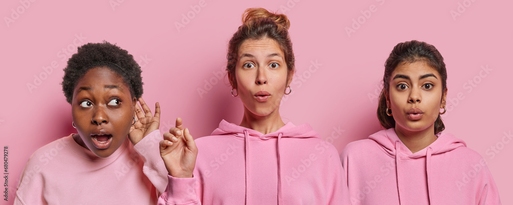 Image of two stunned young women and one African female model keeps hand near ear tries to overhear something looks curiously aside isolated over pink background. People and emotions concept Image of two stunned young women and one African female model keeps hand near ear tries to overhear something looks curiously aside isolated over pink background. People and emotions concept