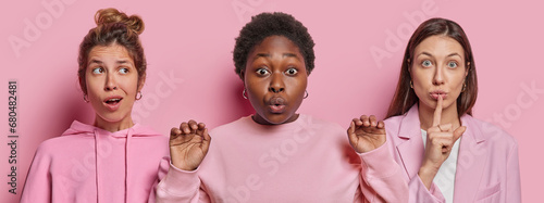 Photo of emotive dark skinned chubby woman stares impressed poses between two women who express secrecy and surprisement dressed in pink clothing stand next to each other. Human reactions concept photo