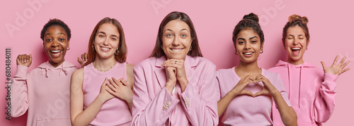 Horizontal shot of five women have broad smiles gesture actively express gratitude and shows heart gesture wear pink casual clothing stand next to each other at studio. Human emotions concept
