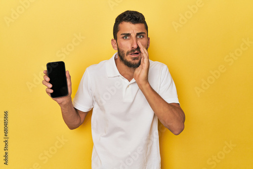 Young Hispanic man shows phone screen is saying a secret hot braking news and looking aside