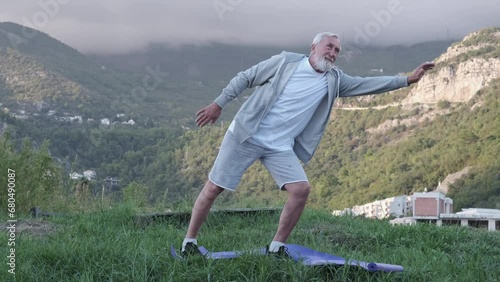 An elderly, gray-haired man with a gray beard in a gray tracksuit practicing yoga against the backdrop of beautiful mountains