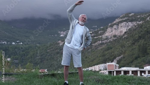 An elderly, gray-haired man with a gray beard in a gray tracksuit practicing yoga against the backdrop of beautiful mountains
