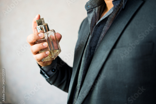 Handsome man in a suit applying perfume on neck against light background at home. Male holding up bottle of luxury perfume. Men perfume photo