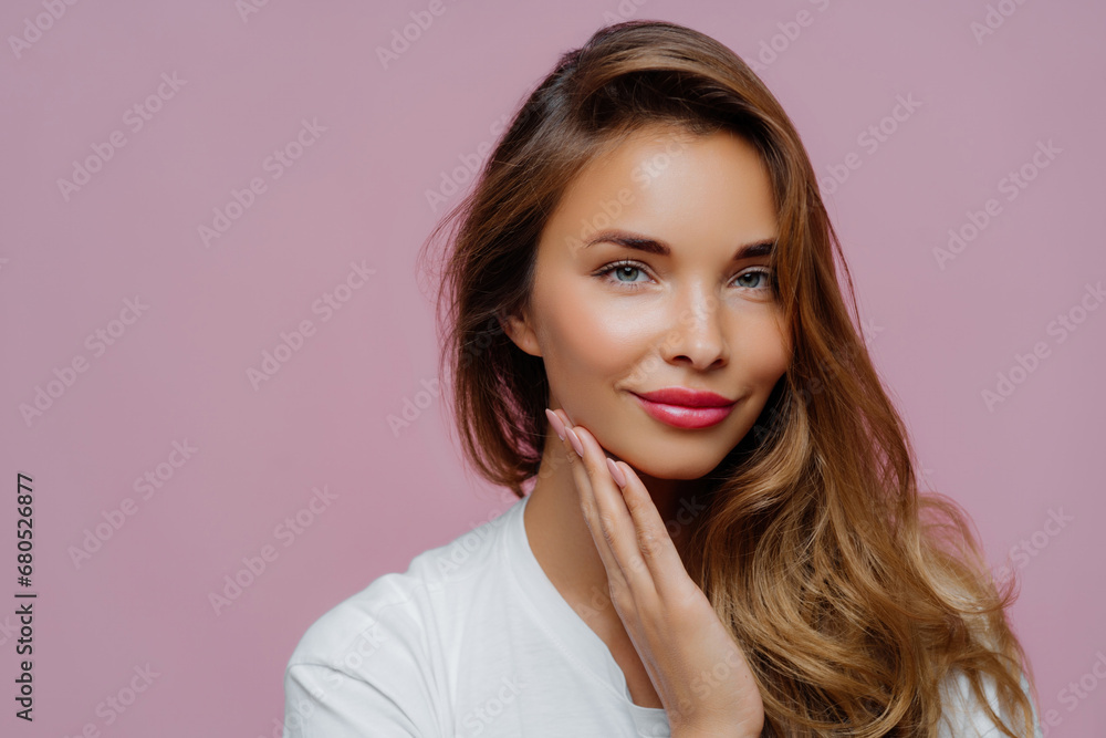 Radiant beauty with hand on cheek and glossy hair, wearing a white shirt, against a pink background Radiant beauty with hand on cheek and glossy hair, wearing a white shirt, against a pink background
