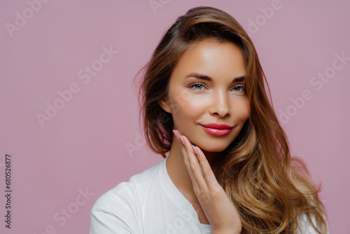 Radiant beauty with hand on cheek and glossy hair, wearing a white shirt, against a pink background