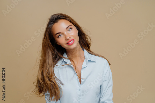 Charming woman with tousled hair and open light blue shirt gazes softly on a beige backdrop. © VK Studio