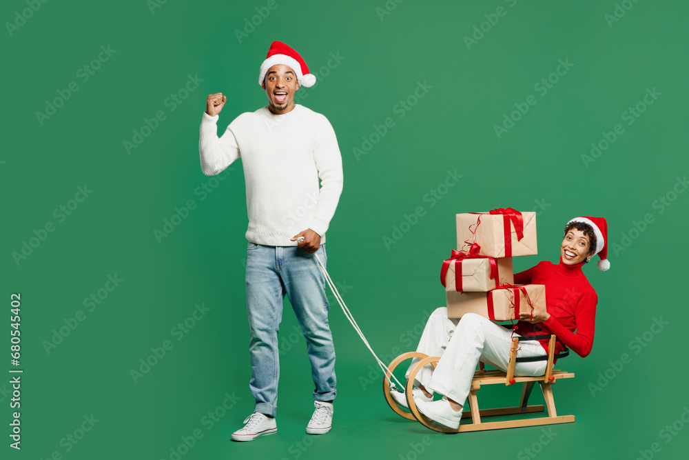 Φωτογραφία Full body merry young couple man woman wear red casual clothes Santa hat posing ride carry on sled hold present box isolated on plain green background