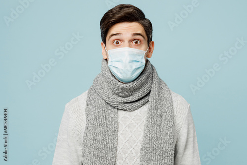 Young surprised ill sick man wear gray sweater scarf sterile mask look camera isolated on plain blue background studio portrait. Healthy lifestyle disease virus treatment cold season recovery concept. photo