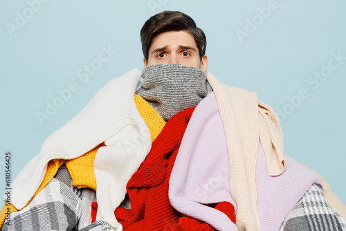 Murais de parede Full body sad frozen young ill sick man wrapped in pile of many clothes looking camera isolated on plain blue background studio