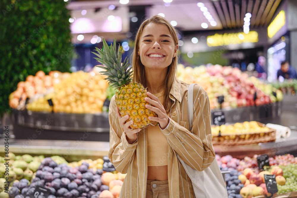 Young smiling happy customer woman wears casual clothes hold pine-apple shopping at supermaket store grocery shop buying choose fruit products inside hypermarket. Purchasing food gastronomy concept.