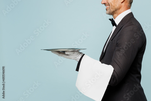 Cropped side view adult barista male waiter butler man wear shirt black suit bow tie uniform hold in hand metal tray plate work at cafe isolated on plain blue background. Restaurant employee concept.
