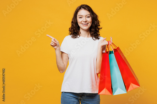 Young smiling woman wears t-shirt casual clothes hold in hand paper package bags after shopping point aside isolated on plain yellow orange color background studio. Black Friday sale buy day concept.