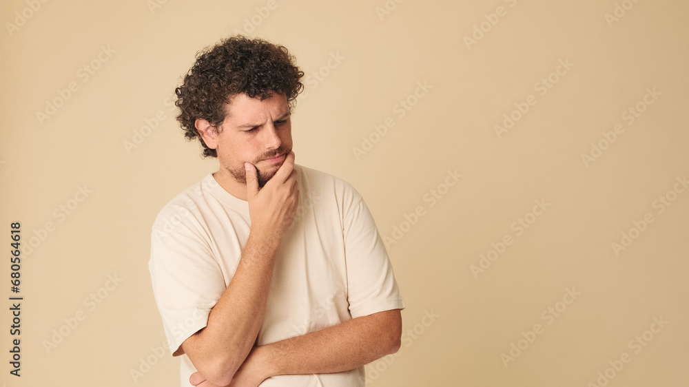 Thoughtful guy with curly hair dressed in beige t-shirt, with his hands folded unable to make up his mind in studio on beige background in the studio Thoughtful guy with curly hair dressed in beige t-shirt, with his hands folded unable to make up his mind in studio on beige background in the studio