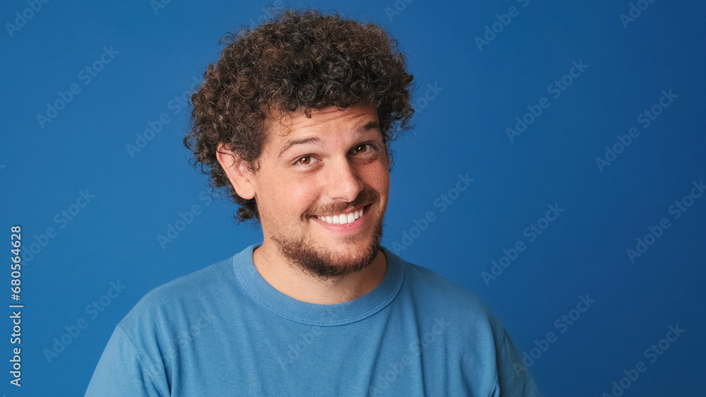 Close up, pensive guy with curly hair dressed in blue t-shirt looks at the camera having good idea on blue background in the studio Close up, pensive guy with curly hair dressed in blue t-shirt looks at the camera having good idea on blue background in the studio