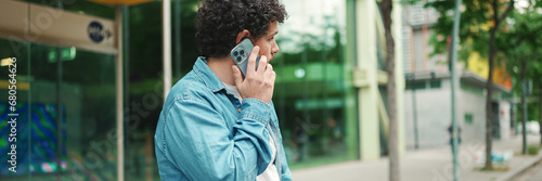 young bearded man in denim shirt stands at bus stop waiting for bus and talking on mobile phone on modern city background