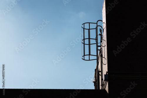 Ladder with safety cage at the factory plant rooftop on sunny day blue sky as background. Building exterior equipment object. Photo contained high contrast ratio between bright and shadow.